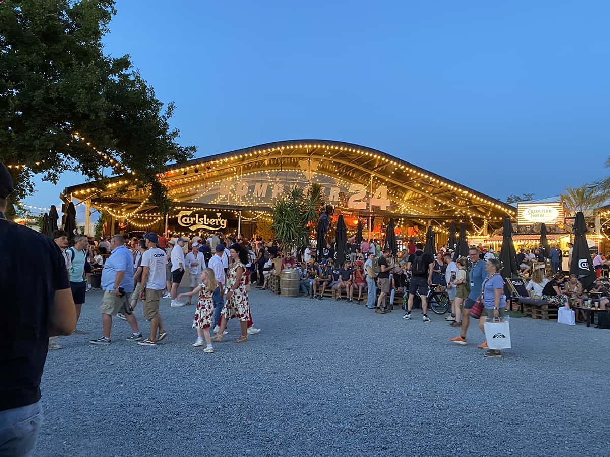 Grande halle Carlsberg Garage 24 illuminée, foule festive et musique DJ au Mans Classic 2025.