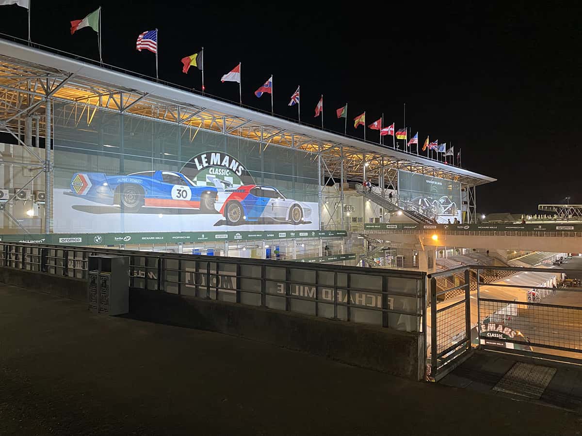 Bâtiment des tribunes décoré « Le Mans Classic » et drapeaux internationaux flottant sous les projecteurs nocturnes.
