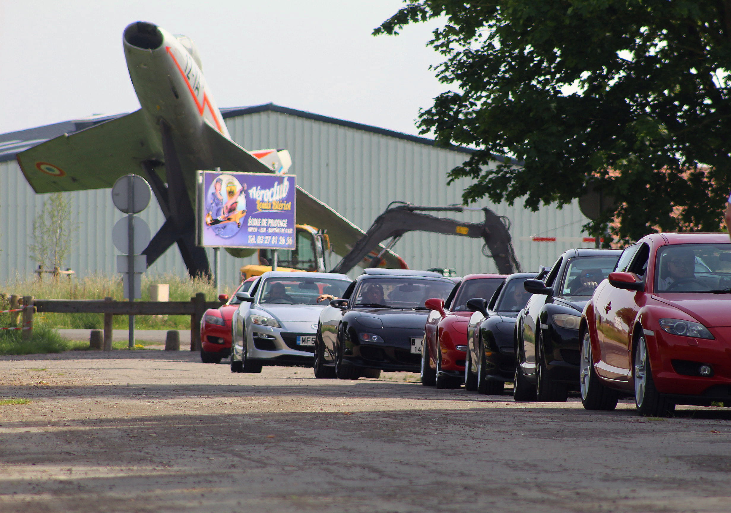 Classic Rotary Day 2026 - le convoi rotatif passe devant l'aérodrome de Niergnies et son avion de chasse Convoi de Mazda RX-8 et RX-7 alignées devant l'aérodrome de Niergnies lors du Classic Rotary Day 2026 - avion de chasse exposé et panneau de l'Aéroclub Louis Blériot en arrière-plan