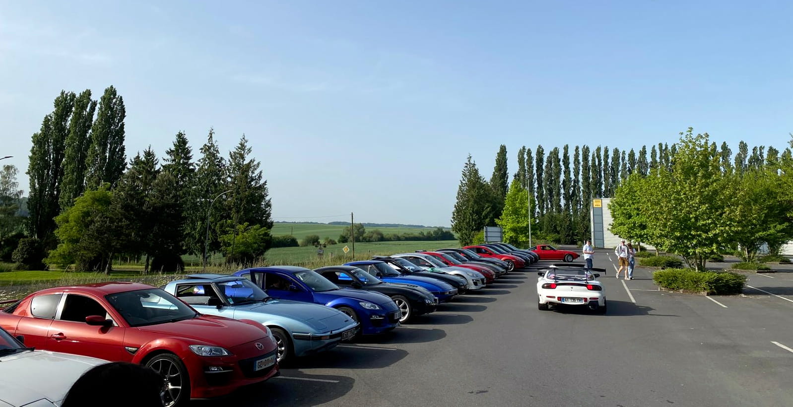 Départ CRD 2026 — parking Action de Bohain-en-Vermandois, Cambrésis Point de départ du Classic Rotary Day 2026 : une vingtaine de Mazda à moteur rotatif alignées dans le parking Action de Bohain-en-Vermandois, Cambrésis — RX-8, RX-7 FD et autres modèles Wankel sous un ciel bleu matinal
