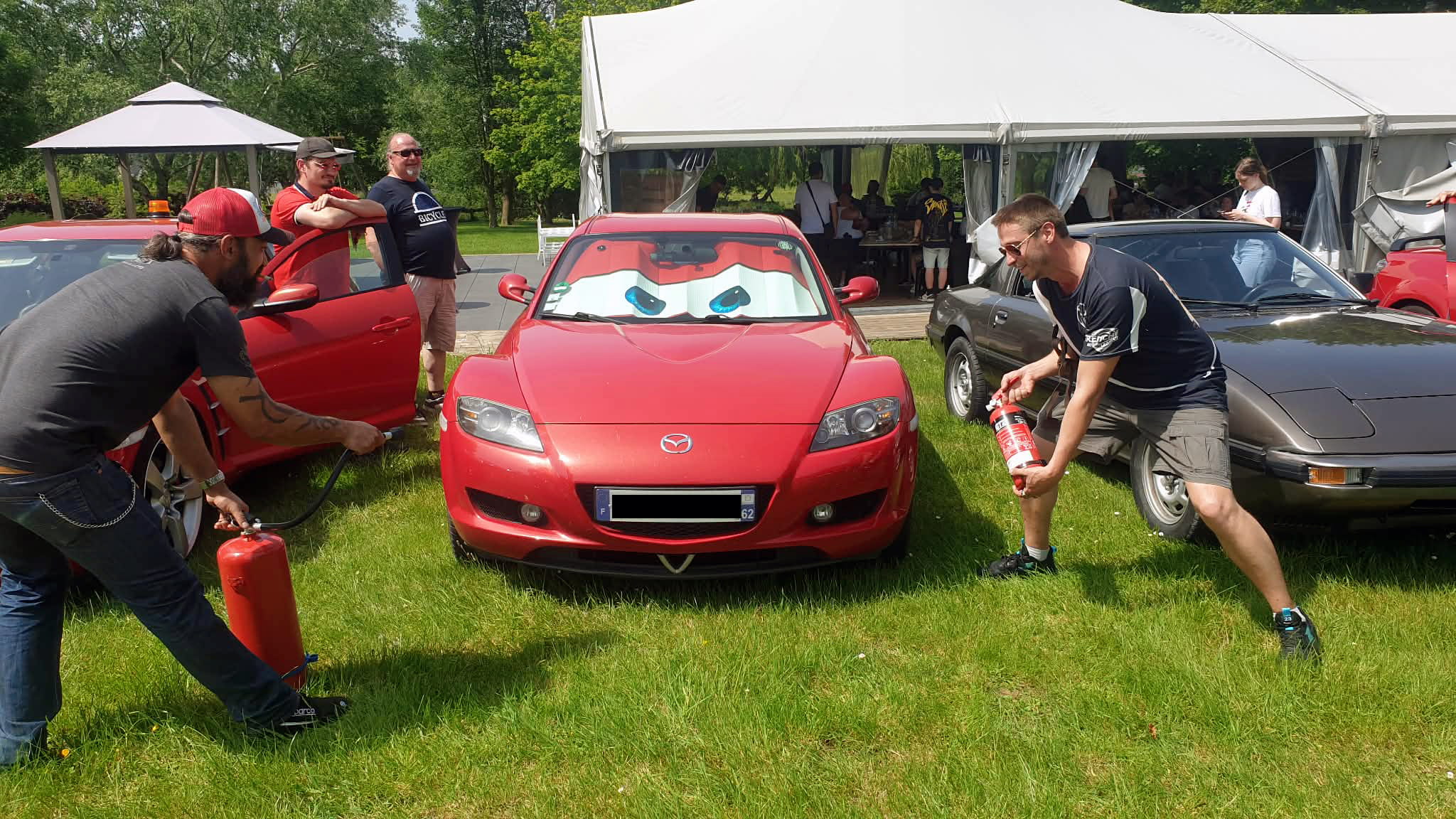 Classic Rotary Day 2026 - quand les propriétaires de rotatif voyagent toujours avec un extincteur 😄 Blague d'initiés au Classic Rotary Day - deux participants s'approchent en souriant d'une Mazda RX-8 rouge avec extincteurs, clin d'œil humoristique à la réputation thermique du moteur Wankel