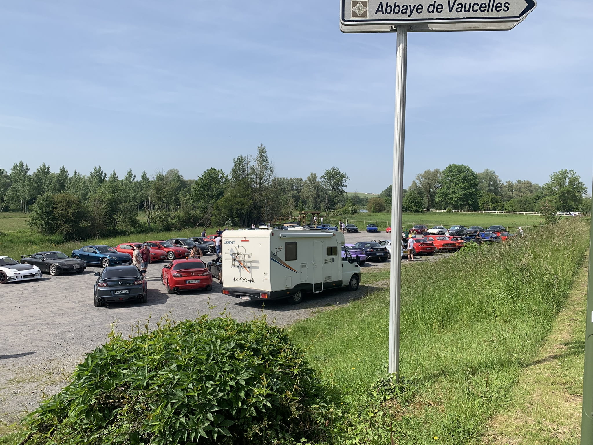 Classic Rotary Day 2026 - rassemblement complet des véhicules sur le parking de l'abbaye de Vaucelles avant le trajet 3 Vue d'ensemble du parking de l'abbaye de Vaucelles avec la totalité des véhicules rotatifs du Classic Rotary Day avant le départ du trajet 3 - panneau directionnel Abbaye de Vaucelles au premier plan, dizaines de Mazda RX-7 et RX-8 garées sur le parking en gravier