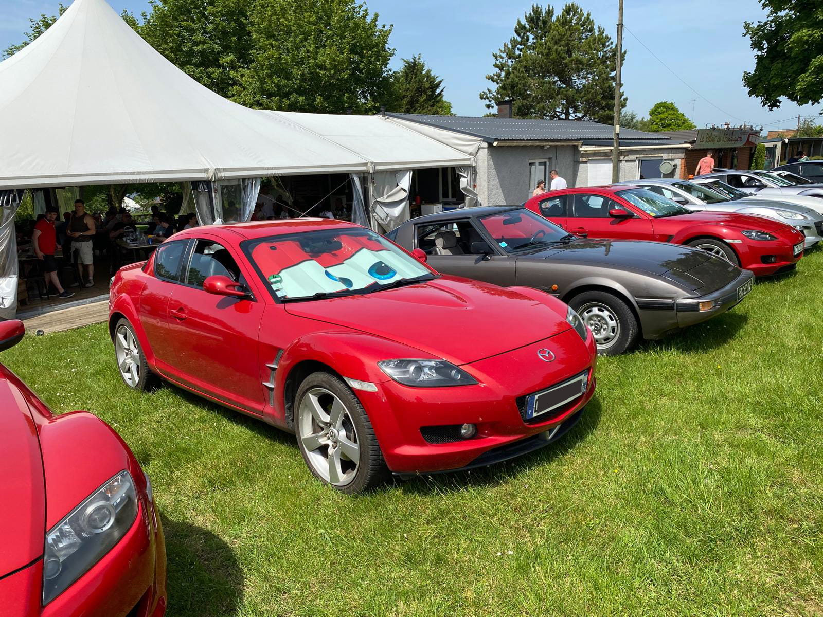 Parking du restaurant Le Relais de l'&Eacute;chauguette lors du Classic Rotary Day - rang&eacute;e de Mazda RX-8 gar&eacute;es sur la pelouse dont une rouge au premier plan, chapiteau et membres du club en arri&egrave;re-plan
