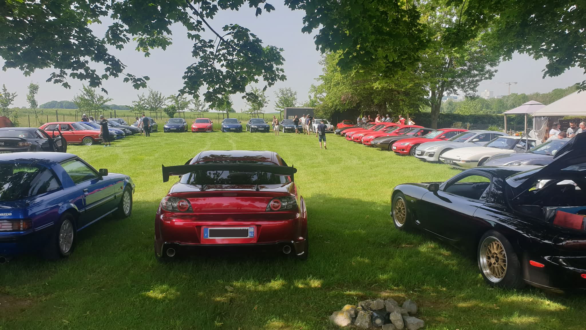 Rassemblement Mazda moteur rotatif - Classic Rotary Day 2026 Vue panoramique du parking pelouse du Classic Rotary Day au Relais de l'Échauguette - dizaines de véhicules à moteur rotatif Mazda alignés des deux côtés, RX-8 rouge préparée avec aileron au centre, RX-7 FD3S noire et Mazda bleue au premier plan