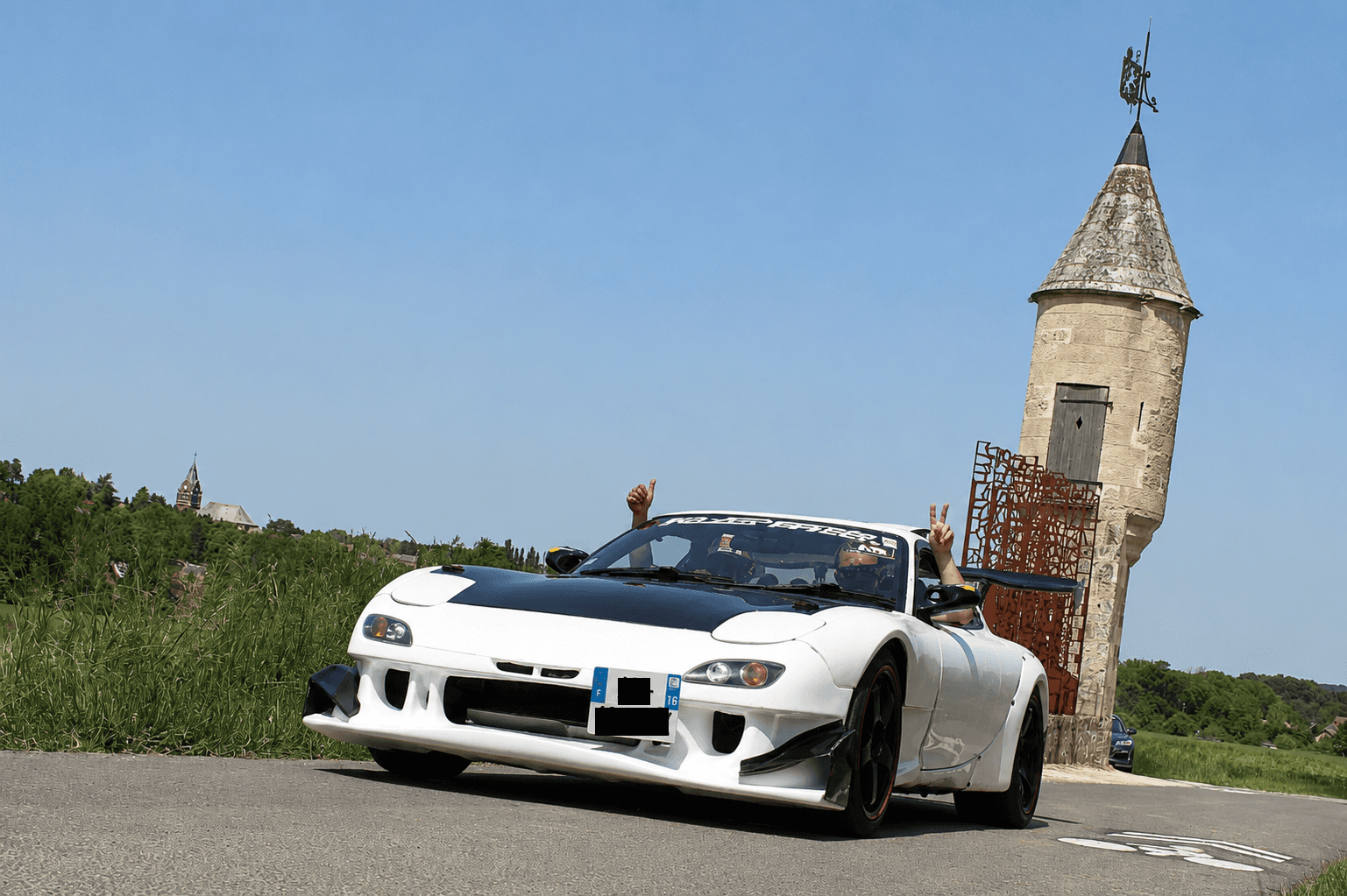Classic Rotary Day 2026 - RX-7 FD3S préparée devant l'échauguette Mazda RX-7 FD3S blanche préparée wide body devant l'échauguette historique lors du Classic Rotary Day - occupants faisant signe à l'objectif, pare-brise frappé du logo RX8-Passion