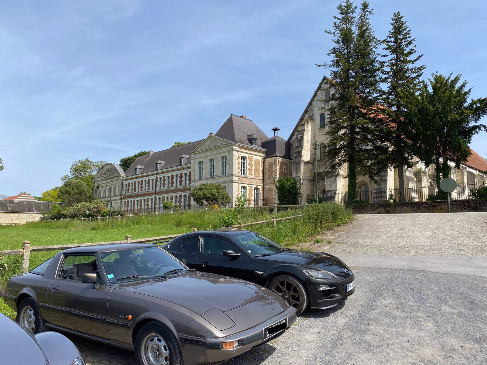 Classic Rotary Day 2026 - stationnement des véhicules rotatifs devant l'abbaye de Vaucelles Mazda RX-7 SA22 bronze et Mazda RX-8 noire stationnées sur le parking de l'abbaye de Vaucelles lors du Classic Rotary Day - façade historique de l'abbaye cistercienne en arrière-plan