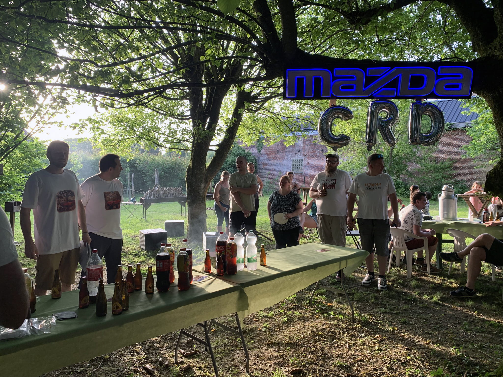 Classic Rotary Day 2026 - la soirée barbecue et le moment de remerciements dans le corps de ferme Soirée barbecue du Classic Rotary Day 2026 dans un corps de ferme - participants réunis autour des tables sous les arbres, enseigne lumineuse Mazda et lettres ballons CRD, ambiance conviviale de fin de journée dans l'Aisne