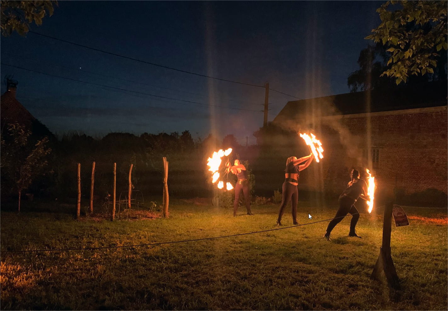 Classic Rotary Day 2026 - le spectacle de feu de clôture à la tombée de la nuit Spectacle de feu nocturne clôturant le Classic Rotary Day 2026 - trois artistes cracheurs et jongleurs de feu en représentation dans le jardin du corps de ferme à la tombée de la nuit, flammes illuminant la scène